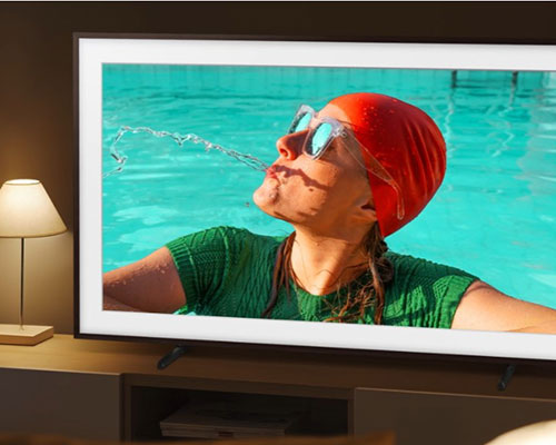 The Frame TV is set in a room with dim lighting, but its screen displays bright, vivid colors powered by Quantum Dot. A woman is spitting out water in the pool. The brightness and 100% color volume powered by Quantum Dot logo is on the lower right side.